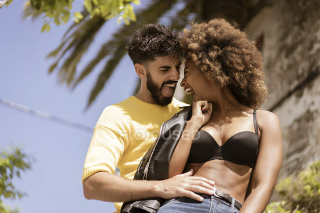 Guapo barbudo chico sonriendo y coqueteando con atractiva mujer negra en sujetador, mientras que de pie en la calle de la ciudad juntos en el día soleado - foto de stock