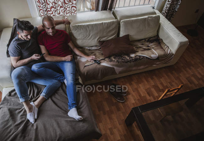 Pensive гей пара зі смартфоном розслабляється на дивані вдома разом — стокове фото