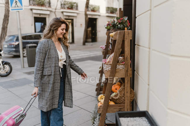 Sonriente mujer joven elegante con maleta mirando los comestibles en los estantes de madera cerca de la tienda en la calle de la ciudad - foto de stock