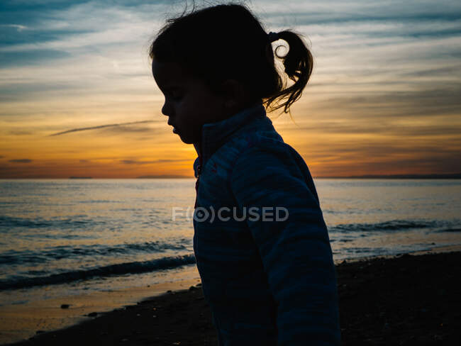 Silueta de niña contra una puesta de sol junto a la orilla del mar - foto de stock