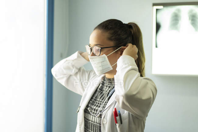 Young female doctor in uniform and medical mask standing in room with x-ray image on wall — Stock Photo