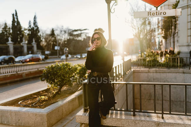 Стильна жінка куріння сигарет недалеко від станції метро на сонячній вулиці — стокове фото
