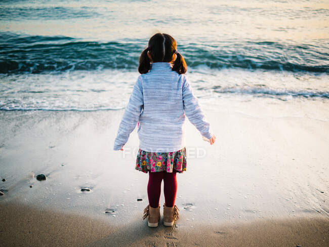 Анонімна маленька дівчинка позаду стоїть на пляжі на піску, дивлячись на море — стокове фото