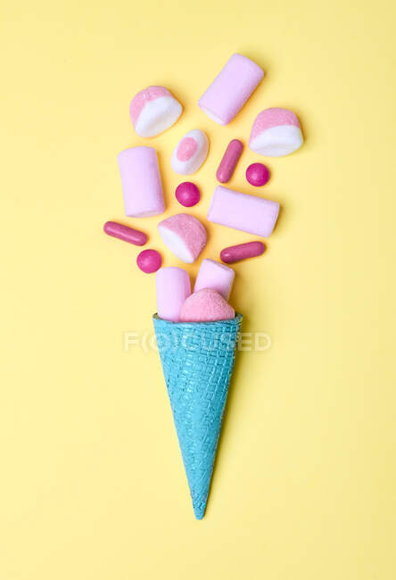 From above of arranged bright blue ice-cream waffle cone with heap of pink candies and marshmallow on yellow background — Stock Photo