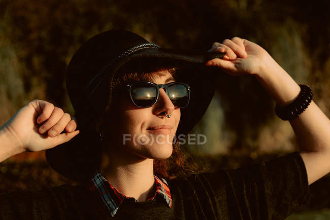 Young sensual brunette wearing black hat and accessories posing in sunlight with sunglasses — Stock Photo