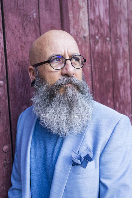 Front view of a mature trendy bearded hipster leaning on a vintage red door outdoors while looking away — Stock Photo