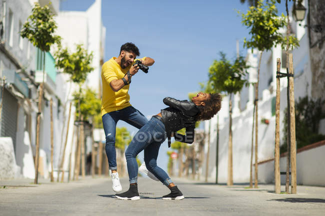 Guapo barbudo chico usando la cámara de fotos para tomar la foto de la mujer gritando y doblando de nuevo en la calle de la ciudad - foto de stock
