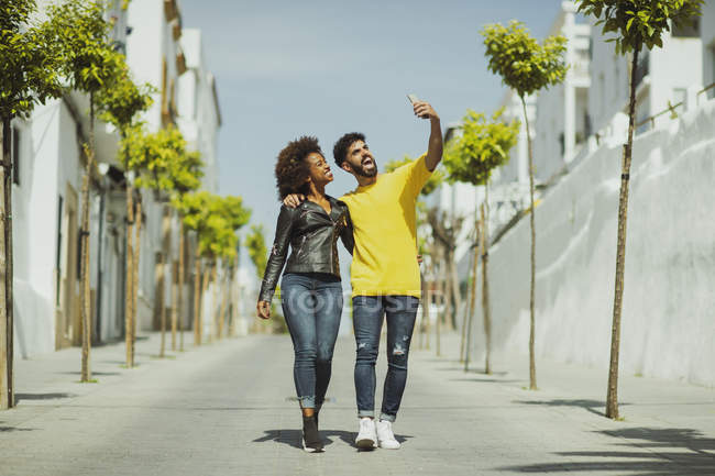 Handsome bearded man and pretty woman walking while taking selfie on sunny day on city street — Stock Photo