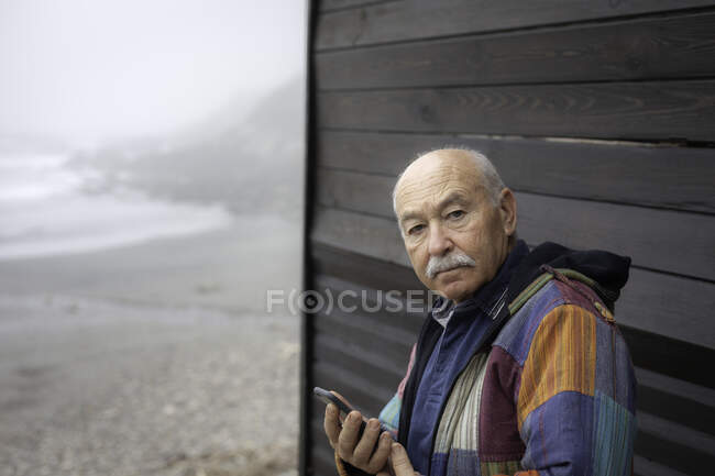 У віці серйозний чоловік в куртці тримає смартфон, дивлячись на камеру проти дерев'яного будинку на віддаленому узбережжі — стокове фото