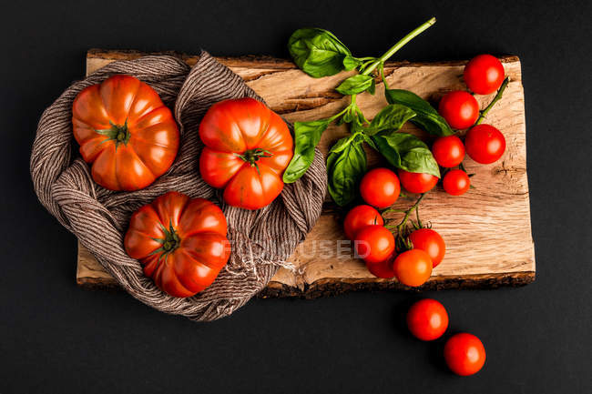 Tomates frescos maduros y hojas de albahaca sobre un trozo de madera sobre fondo negro - foto de stock