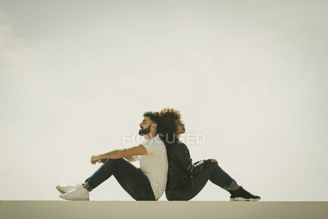 Giovane uomo e donna multietnici seduti schiena a schiena sul muro di costruzione contro il cielo grigio — Foto stock