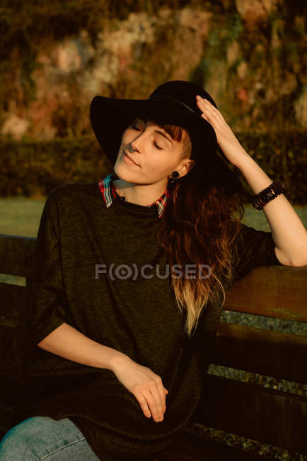 Молода чуттєва брюнетка в чорній шапці та аксесуарах сидить на лавці на сонячному світлі — стокове фото