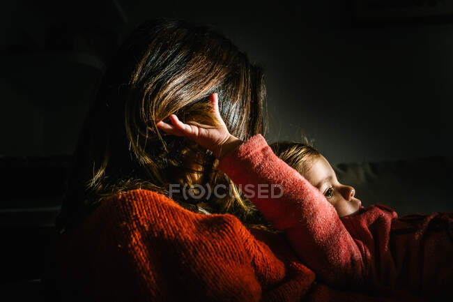 Dentro de casa cena de uma mãe anônima abraçando sua filha no sofá sob iluminação dramática — Fotografia de Stock