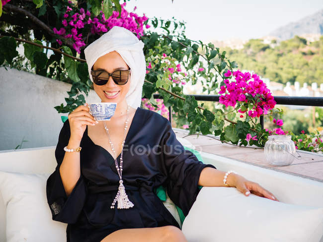 Mulher em vestido de cetim preto e toalha na cabeça descansando em uma varanda desfrutando de belas vistas em uma manhã brilhante ensolarada — Fotografia de Stock