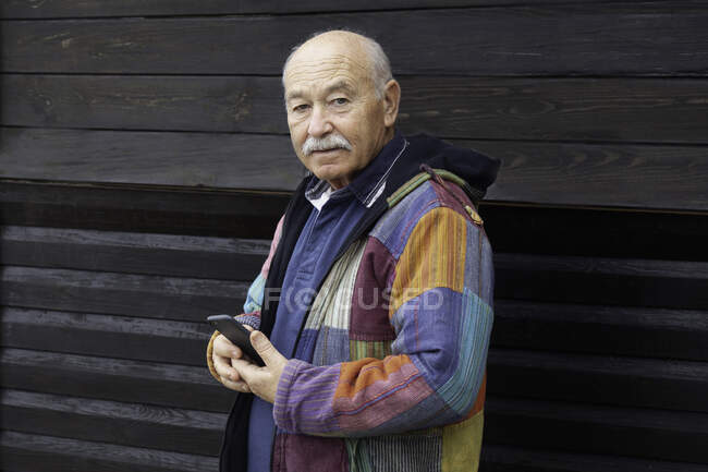 У віці серйозний чоловік в куртці тримає смартфон, дивлячись на камеру проти дерев'яного будинку — стокове фото