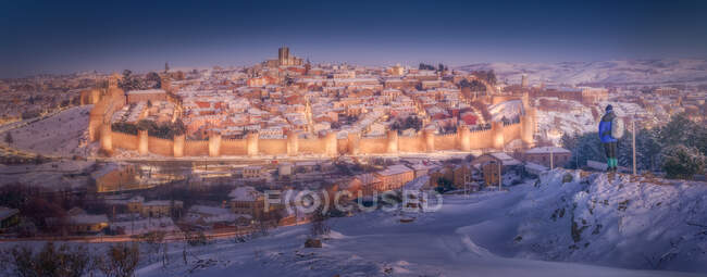 Vista trasera del viajero admirando pintoresca vista de la ciudad iluminada nevada contra el crepúsculo cielo fondo - foto de stock