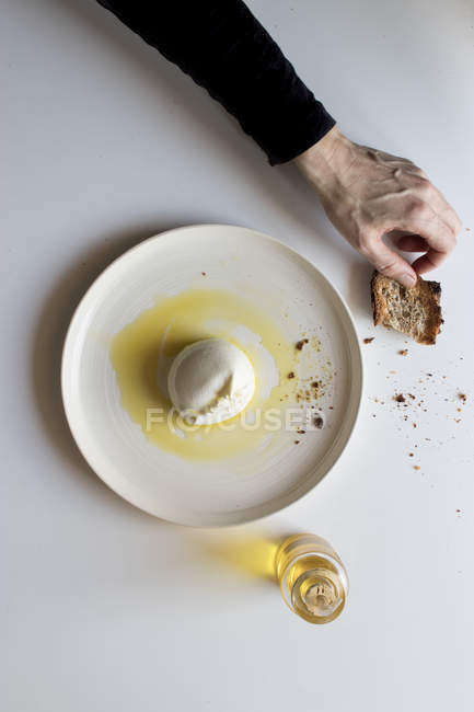 Mano de persona anónima sosteniendo pedazo de pan cerca con sabrosa burrata con aceite sobre fondo blanco - foto de stock