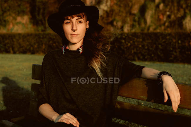 Junge sinnliche Brünette mit schwarzem Hut und Accessoires sitzt auf Bank im Sonnenlicht — Stockfoto