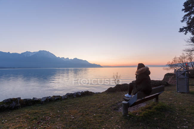 Back view of woman sitting on wood seat above turquoise lake in snowy mountains of Switzerland — Stock Photo