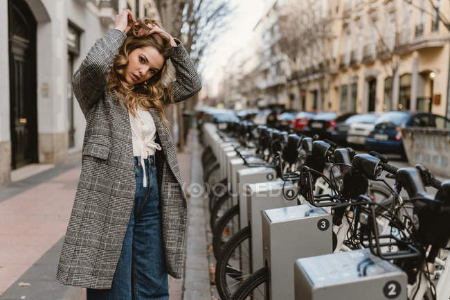 Elegante joven dama de pie en el estacionamiento de bicicletas de alquiler en la ciudad - foto de stock