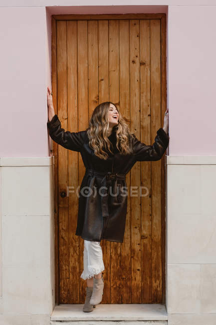 Улыбающаяся стильная женщина в винтажном кожаном пальто, стоящая возле деревянной двери на улице — стоковое фото
