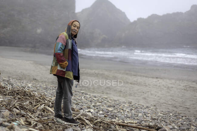 Обличчя старця в пальто, що стоїть на безлюдному узбережжі океану, дивлячись на камеру — стокове фото