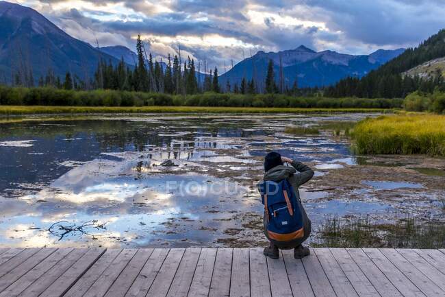 Back view of tourist with backpack shooting on camera picturesque view of water surface and shore with stone hills and cloudy sky in Banff, Canada - foto de stock
