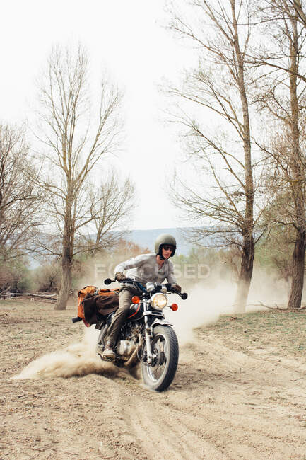Homme en casque à moto rapide sur route de campagne poussiéreuse près des arbres sans feuilles dans la nature — Photo de stock