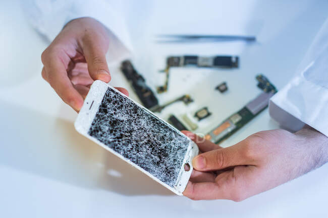 Hands of unrecognizable technician demonstrating cracked screen of modern mobile phone over white table in workshop — Stock Photo