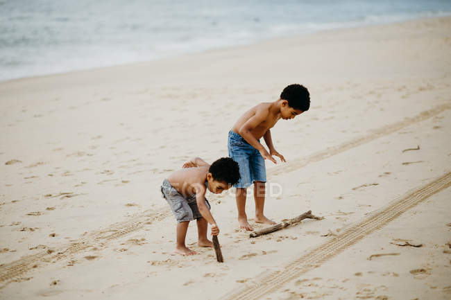 Two African American brothers with sticks playing together on sandy shore near sea — Stock Photo