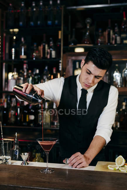 Giovane elegante barman che lavora dietro un bancone del bar versando bevanda da shaker a un bicchiere — Foto stock
