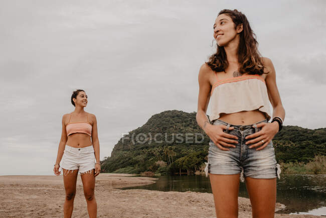 Dos delgadas hembras jóvenes en pantalones cortos y sujetadores sonriendo y mirando a diferentes lados mientras están de pie en la orilla arenosa contra el cielo gris nublado - foto de stock