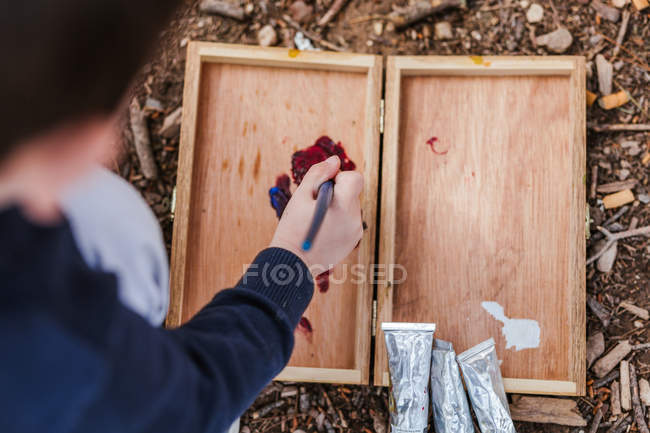Unrecognizable boy mixing red paint on wooden palette while sitting on ground in countryside — Stock Photo