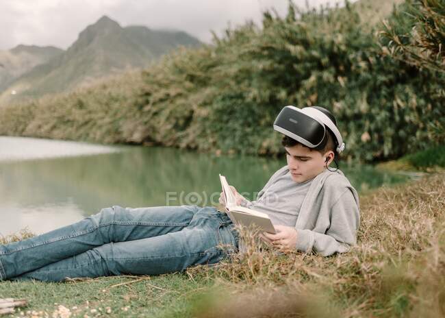 Junger Heranwachsender mit Virtual-Reality-Brille liegt mit Buch im Gras neben einem See — Stockfoto