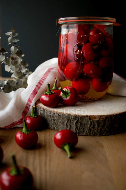 Stillleben mit einem Glas lecker eingelegter Pimientos-Paprika auf einem Stück Holz in der Nähe von Stoffserviette und Pflanzenzweig. — Stockfoto