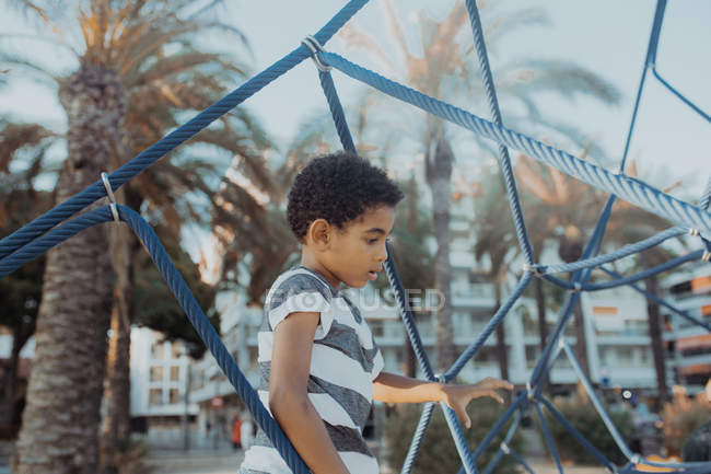 Alegre menino afro-americano rindo e olhando para longe enquanto sentado na rede no playground na praia — Fotografia de Stock