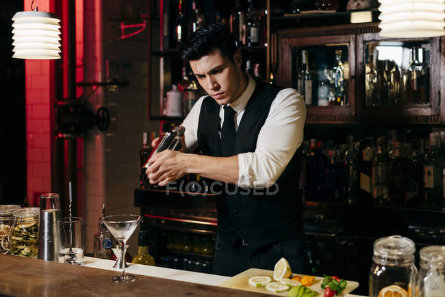 Joven barman elegante que trabaja detrás de un mostrador de bar mezclando bebidas en una coctelera - foto de stock