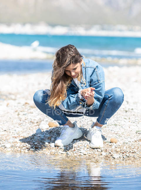 Young woman in denim outfit sitting on haunches on coast and splashing sea water on sunny day — Stock Photo