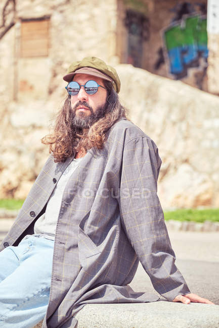 Stylish bearded man with long hair sitting on street with sunglasses — Stock Photo