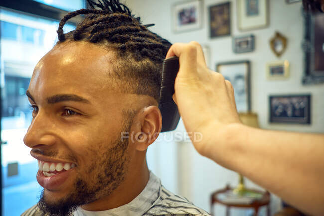 Vista Crop di parrucchiere anonimo facendo un taglio di capelli moderno a un allegro cliente afro-americano — Foto stock