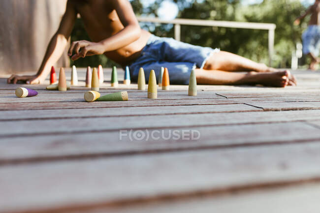 Неузнаваемый афроамериканец, сидящий на деревянной поверхности и играющий с красочными конусами в солнечный день — стоковое фото
