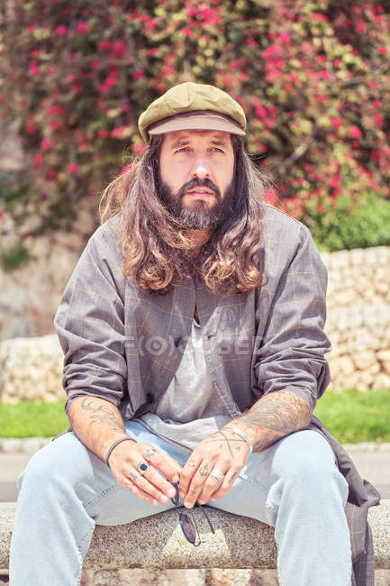 Hombre barbudo con estilo con el pelo largo sentado en el parque y pensando - foto de stock