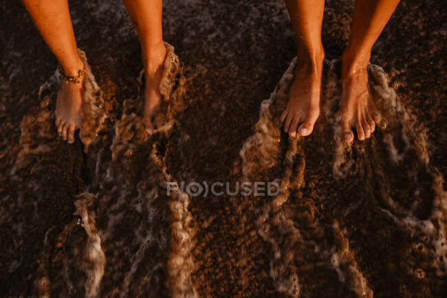 Gambe scalze di donne irriconoscibili in piedi sulla riva bagnata vicino al mare spruzzi in serata — Foto stock