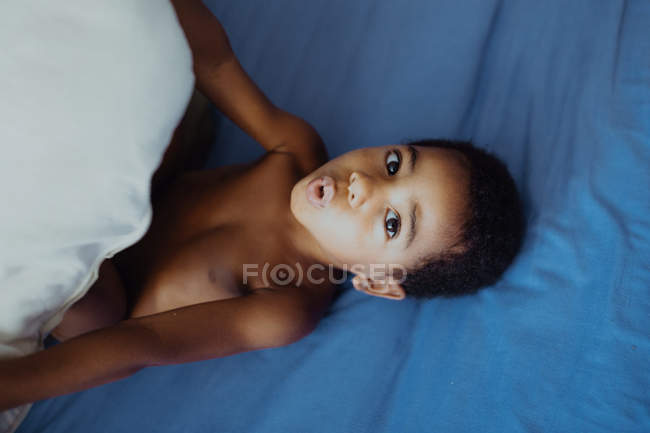 From above adorable African American kid grimacing and holding pillow while playing on comfortable bed at home — Stock Photo