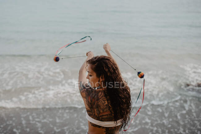 Back view of young tattooed female in swimwear swinging threads with balls while dancing near stormy sea — Stock Photo