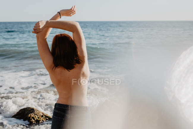 Visão traseira da mulher de topless posando para ondas espumosas de mar tempestuoso no dia ensolarado na natureza — Fotografia de Stock