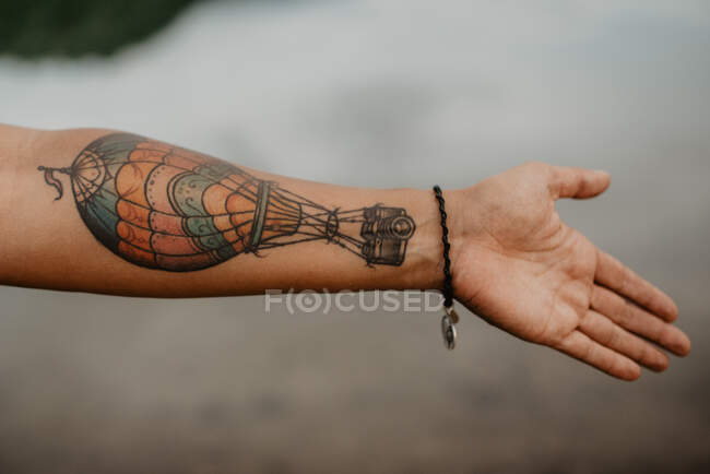 Mulher anônima mostrando braço com tatuagem de balão de ar quente sobre a superfície tranquila do lago no campo — Fotografia de Stock