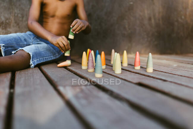 Garçon afro-américain torse nu méconnaissable assis sur une surface en bois et jouant avec des cônes colorés le jour ensoleillé — Photo de stock