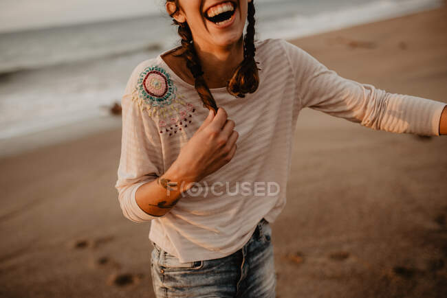 Девушка в повседневной одежде трогает косу и громко смеется, стоя на песчаном берегу. — стоковое фото