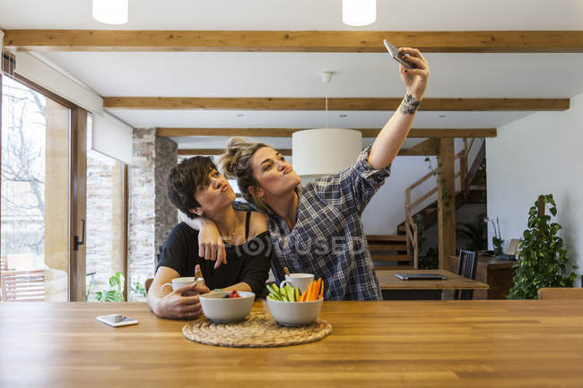 Two beautiful and young women having breakfast at home and having fun, take a auto photo with the smart phone — Stock Photo
